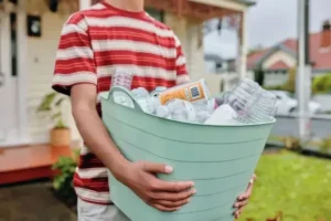 A person in a striped shirt carries a light blue basket filled with empty plastic bottles and containers, standing outdoors near a house with a porch and greenery.