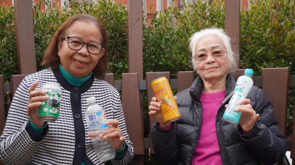 Two people are sitting outdoors holding various beverage containers. One holds a can and a small water bottle, and the other holds two bottles. They are in front of a wooden fence with greenery visible behind them.